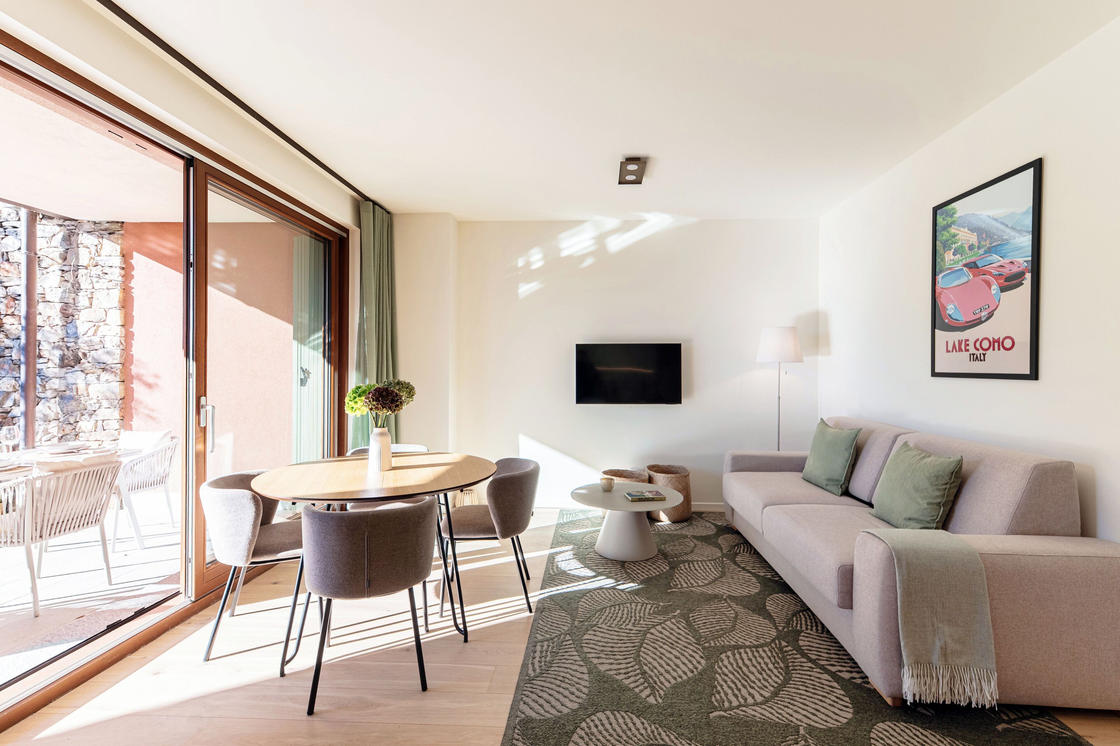 A modern living room with a sofa, a dining table with chairs, a TV mounted on the wall, and a large window with a view of the outdoors.