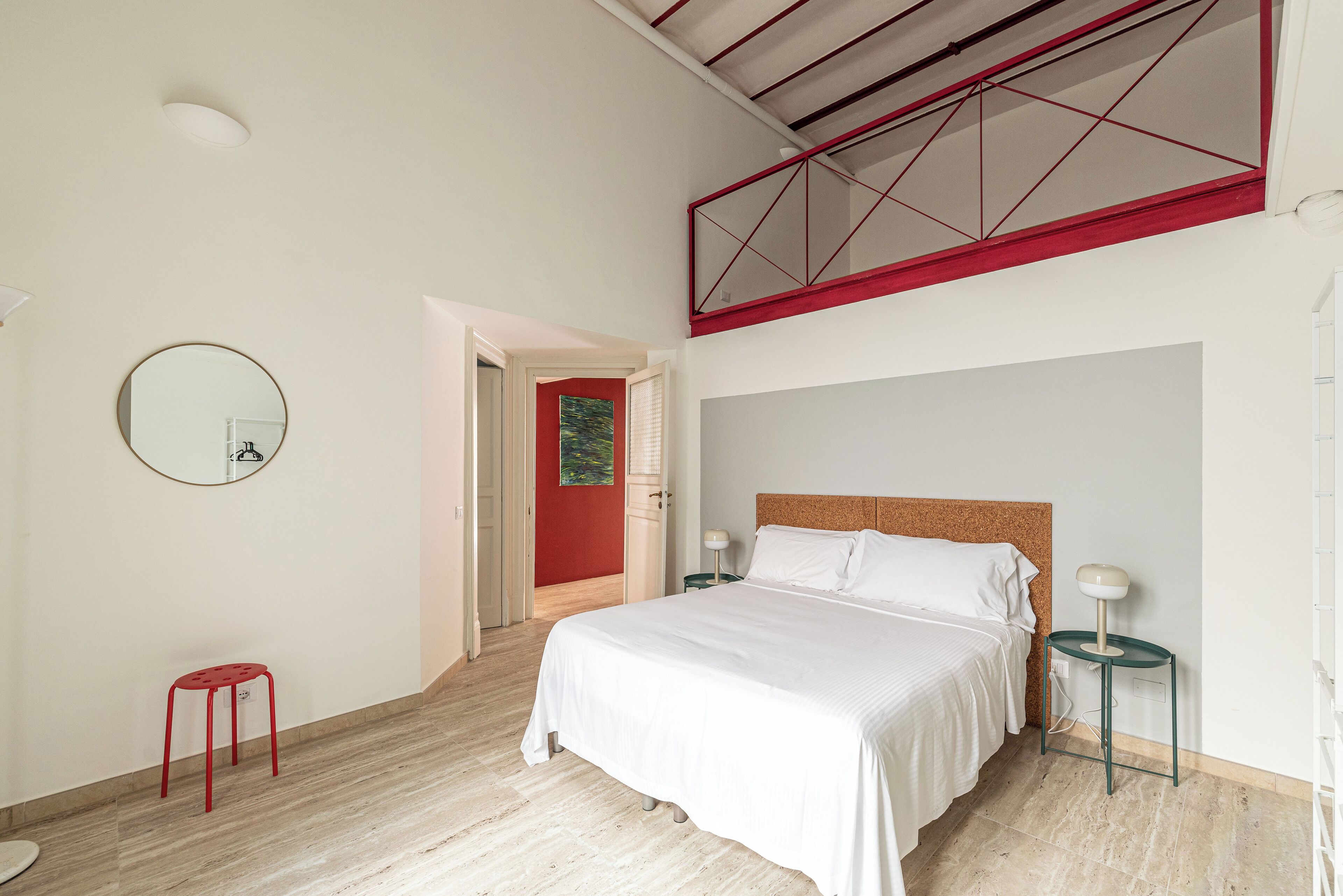 A bedroom with a bed, a red stool, a round mirror, and a door leading to another room.
