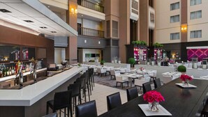 A modern hotel lobby with a bar area, multiple dining tables, and a spacious atrium with a view of the building's exterior.