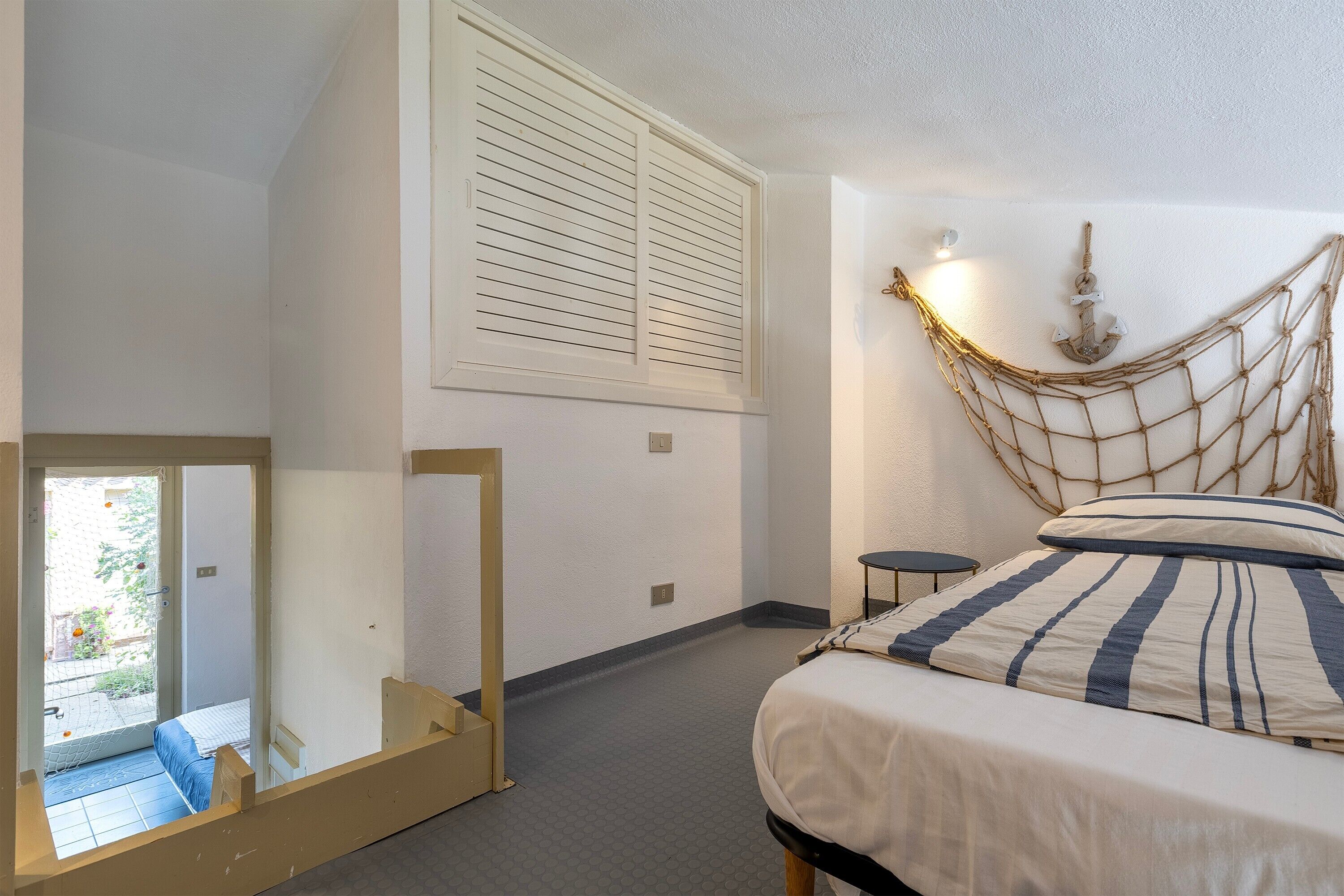 A bedroom with a bed, a small bedside table, a hanging net, and a window with blinds.