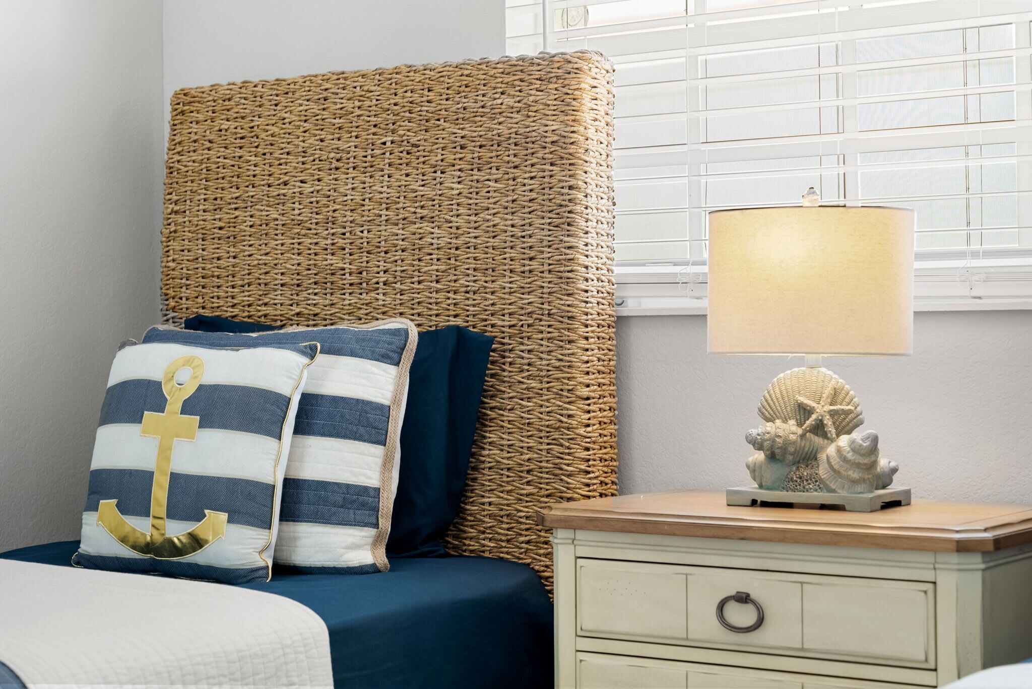 A bedroom with a wicker headboard, striped pillows, a nightstand with a lamp, and a decorative shell.
