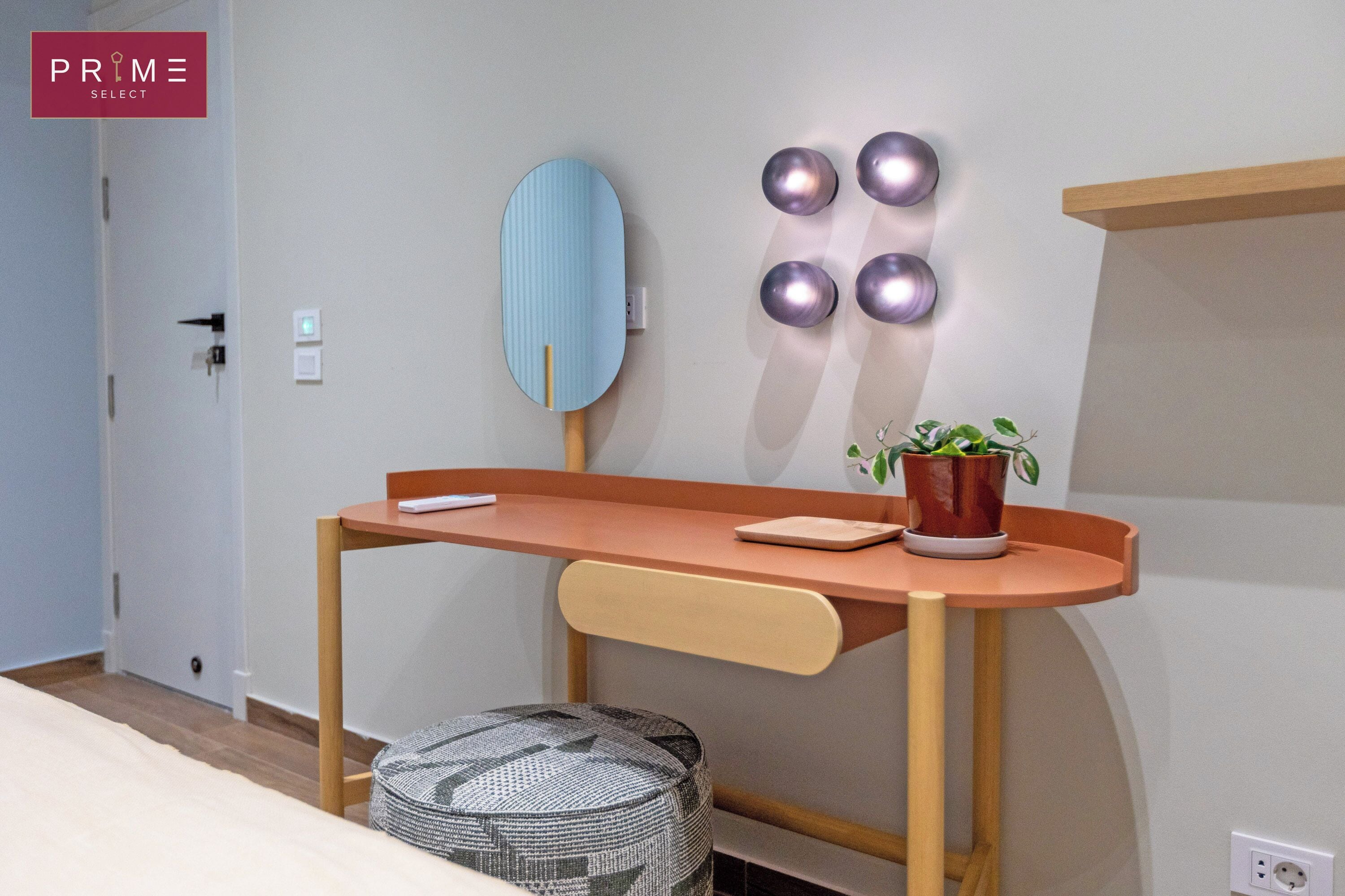 Una habitación moderna con un escritorio de madera, una silla, un pequeño taburete, un espejo adosado a la pared y una planta en maceta.