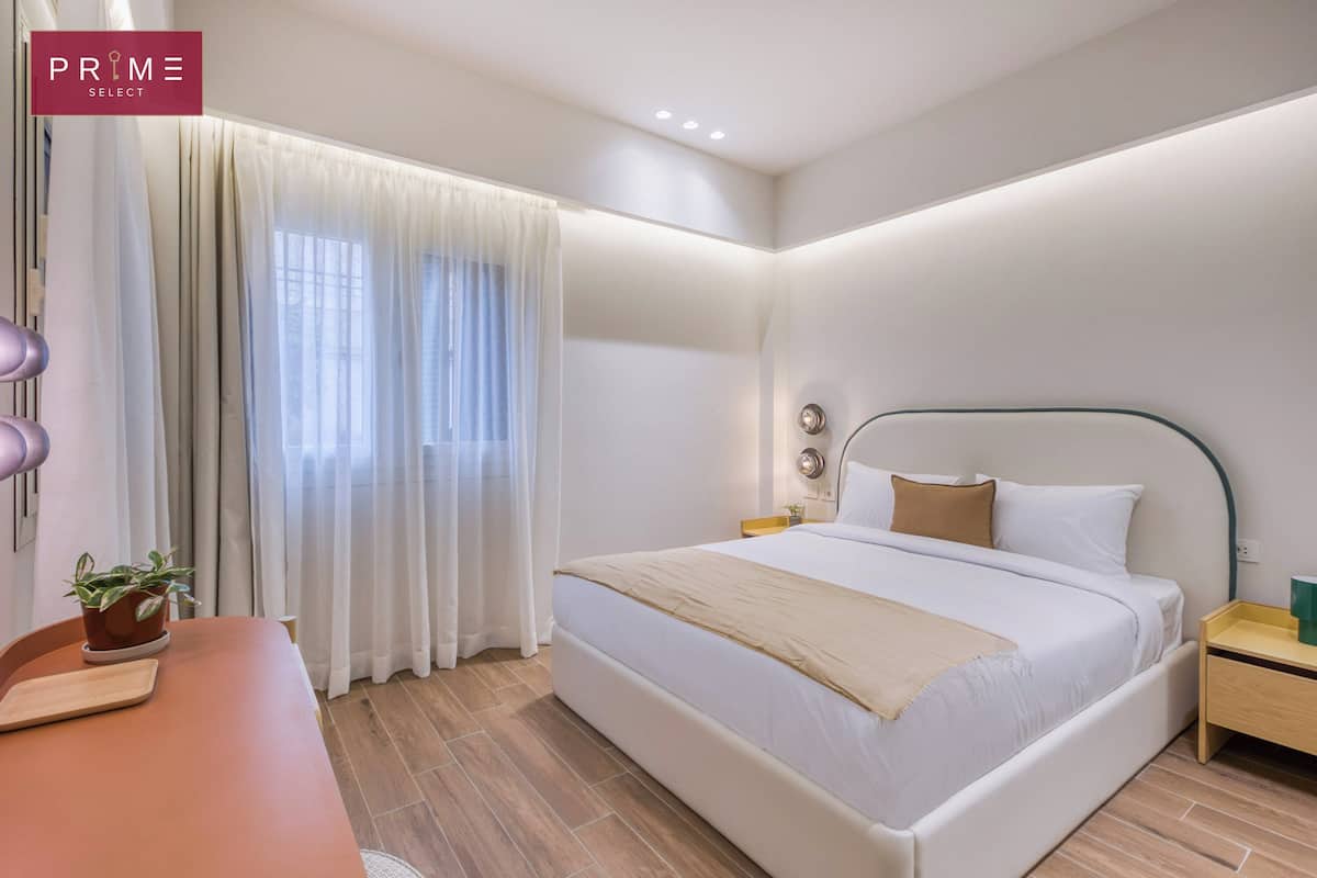 Une chambre d’hôtel moderne avec un grand lit, une table de chevet, une petite table, une plante en pot et une fenêtre avec des rideaux.