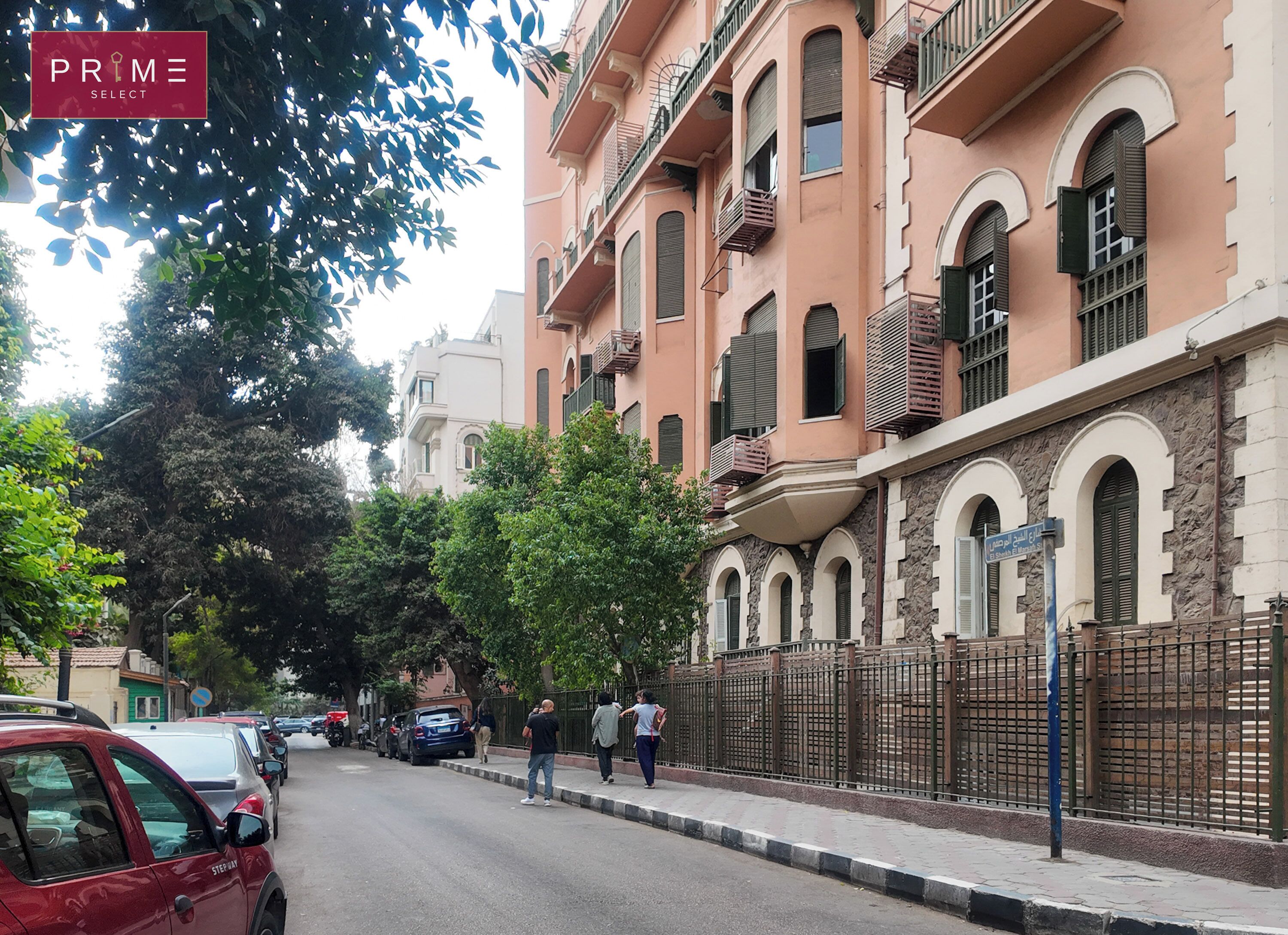 Une rue bordée de voitures garées, avec des piétons et un immeuble doté de balcons et de fenêtres en arc de cercle.