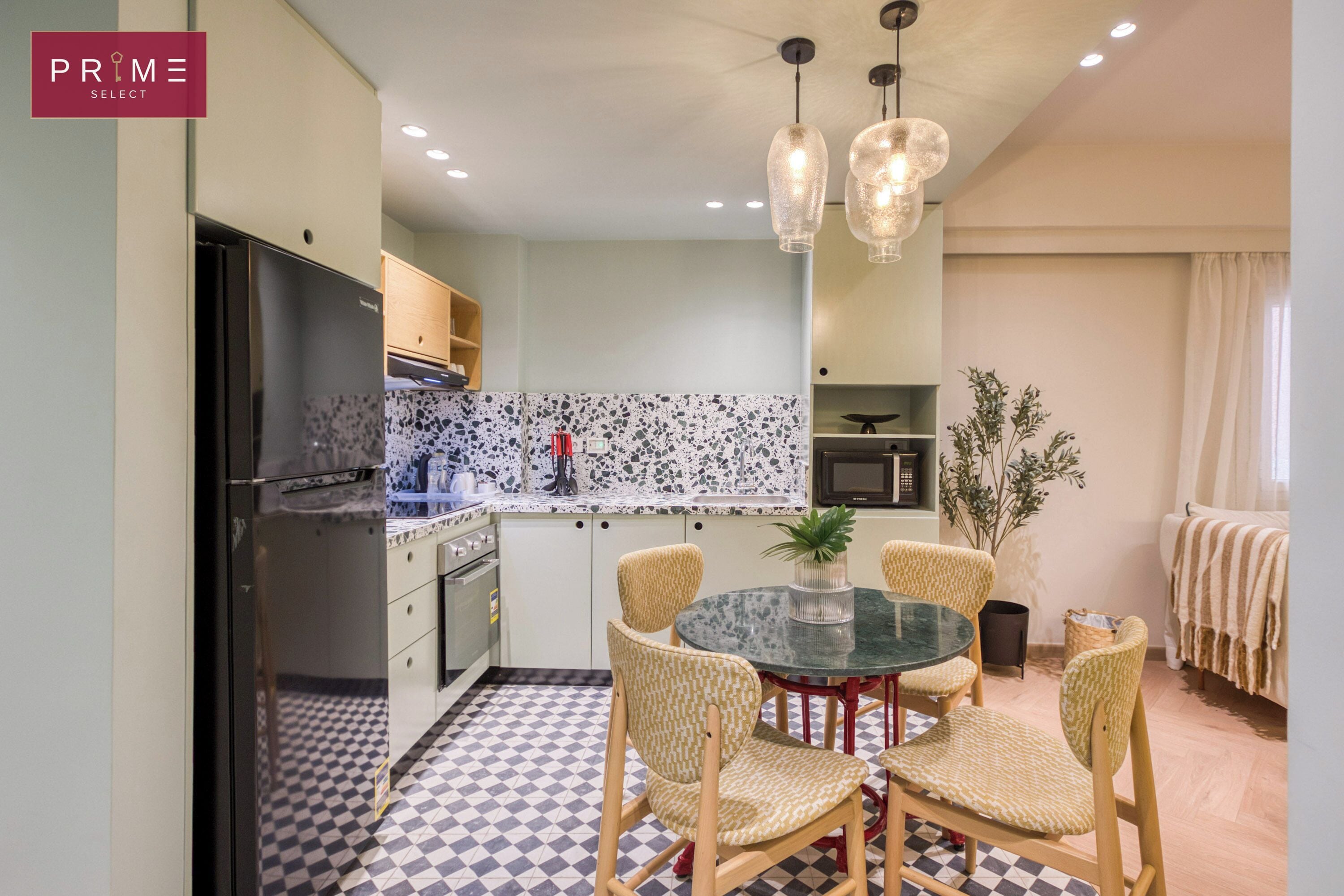 A compact kitchen with a black refrigerator, a microwave, and a stove. There is a dining table with four chairs and a potted plant. The floor has a checkered pattern.