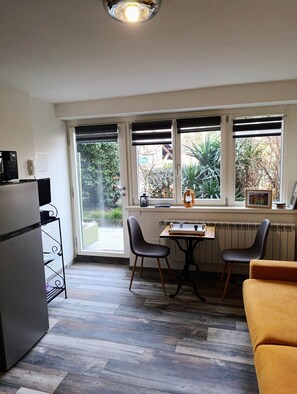 A compact kitchen with a refrigerator, a dining table with two chairs, and a window with a view of greenery. - Altea (Dervio)