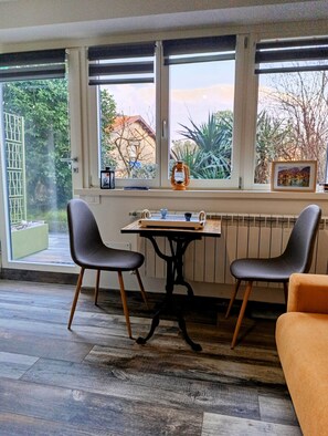 A room with a table and two chairs, a radiator, and a window with a view of trees and a building. - Altea (Dervio)
