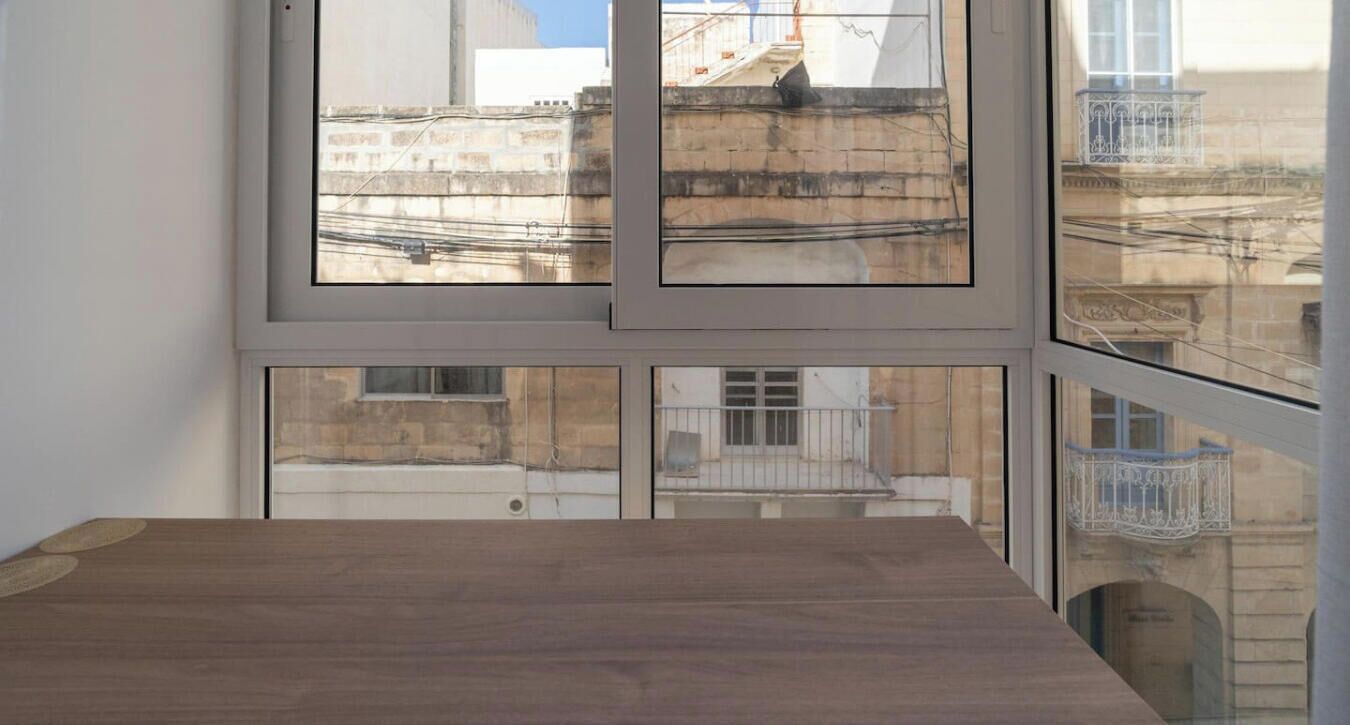 Une pièce dotée d’une grande fenêtre donnant sur un paysage urbain, d’une table en bois et d’une vue sur un immeuble avec un balcon.