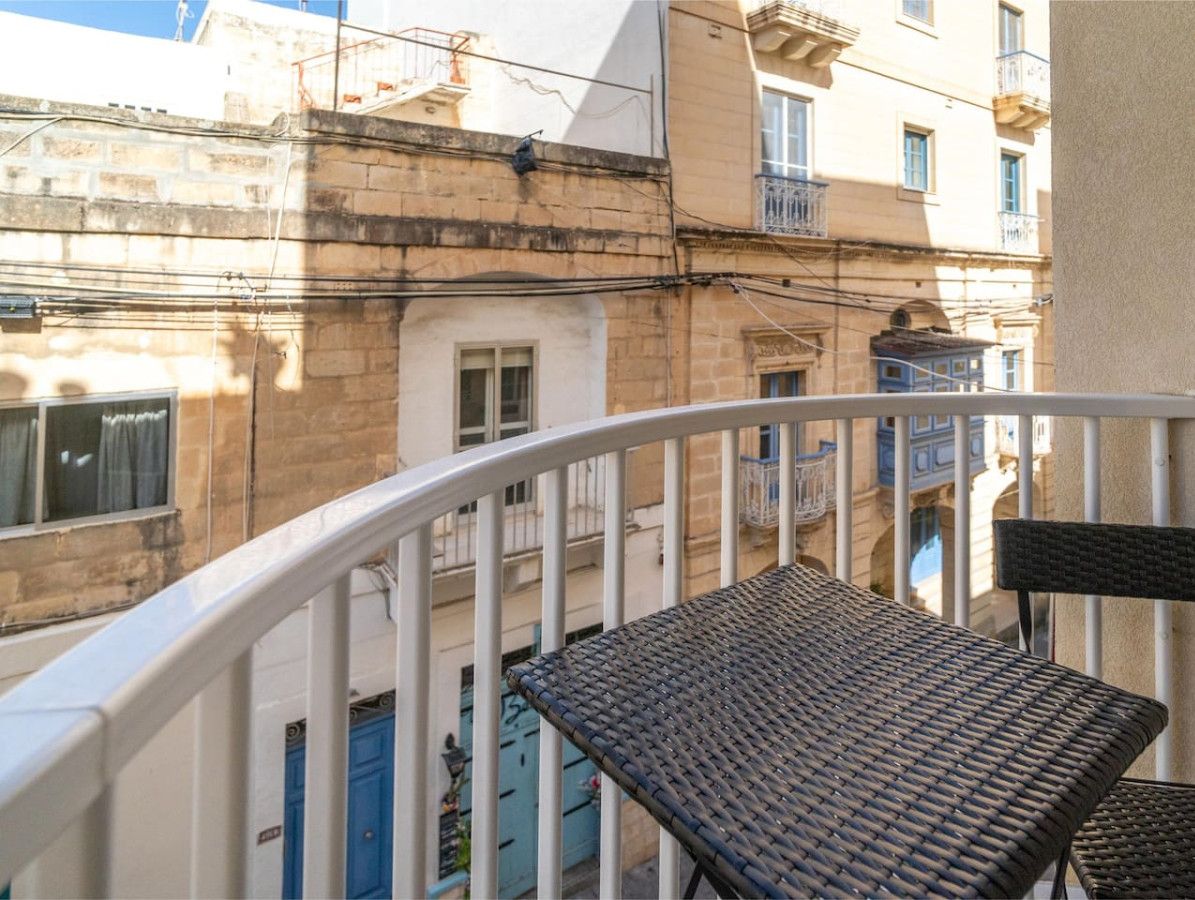 Een balkon met twee rieten stoelen en een witte balustrade, met uitzicht op een gebouw met meerdere ramen en balkons.