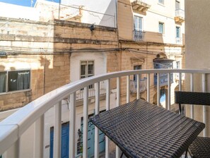 Ein Balkon mit zwei Korbstühlen und einem weißen Geländer, mit Blick auf ein Gebäude mit zahlreichen Fenstern und Balkonen.