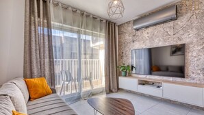 A modern living room with a sofa, a coffee table, a TV, and a balcony with curtains. - Central 2BR Home in Pieta`s Heart w/Wifi & AC ! (Pietà)