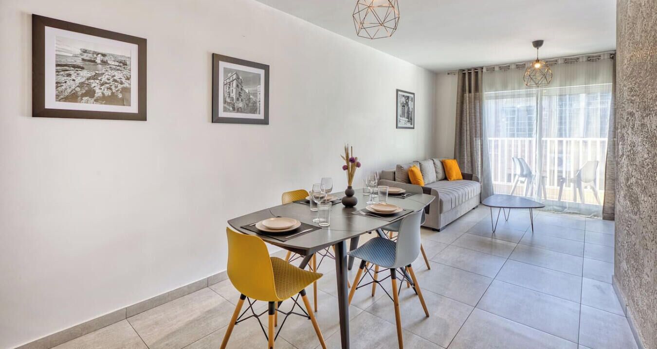 Un comedor con una mesa dispuesta para dos personas, un sofá con cojines y un balcón con cortinas.