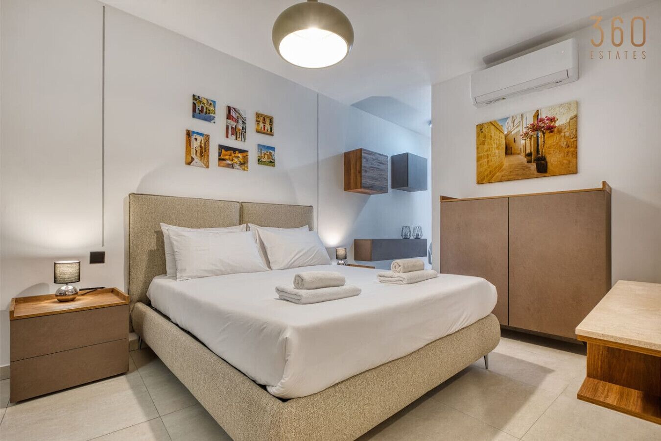 A modern bedroom with a large bed, bedside tables, a wooden nightstand, a headboard, a wall-mounted air conditioner, and artwork on the walls.