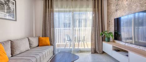 A modern living room with a grey sofa, a wooden coffee table, a TV on a wooden cabinet, and a balcony with a view.