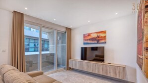 A modern living room with a sofa, a TV mounted on a wooden console, and a large window with curtains.