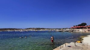 A beach with clear blue water, a sandy shore, and a few people swimming. - Peaceful 2BR Penthouse with Terrace Views & Beach (Marsaskala)