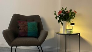 Een moderne fauteuil met een kleurrijk kussen, een klein bijzettafeltje met een bloemenvaas en een kaars, en een vloerbedekking.
