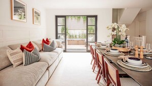 A modern living room with a sofa, dining table, and chairs. There is a large window with blinds, a vase with flowers, and framed pictures on the wall. - Beautiful Fulham Townhouse with a bar & pool table (Parsons Green, London, UK)