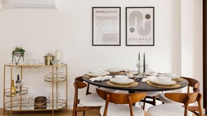 Uma sala de jantar com uma mesa redonda posta para seis pessoas, cercada por cadeiras de madeira com almofadas brancas. Um criado-mudo dourado com copos de vidro está à esquerda. Duas obras de arte emolduradas estão penduradas na parede acima da mesa.