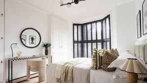 Quarto com cama grande, escrivaninha, cadeira, espelho redondo e janela com persianas.