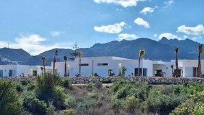 Un edificio moderno con un muro de piedra y palmeras frente a él, con montañas al fondo.