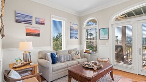 A living room with a beige sofa, a wooden coffee table, and a view of the outdoors through large windows. - Salty Daze- Private pool & hot tub, pet friendly, family friendly! (Cape San Blas)