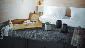 Ein gemütliches Zimmer mit Holzbalkendecke, einem Bett mit weißer Bettwäsche, einer kleinen Holzkiste, einem Topfpflanzen und zwei schwarzen Vasen auf einer gewebten Matte.