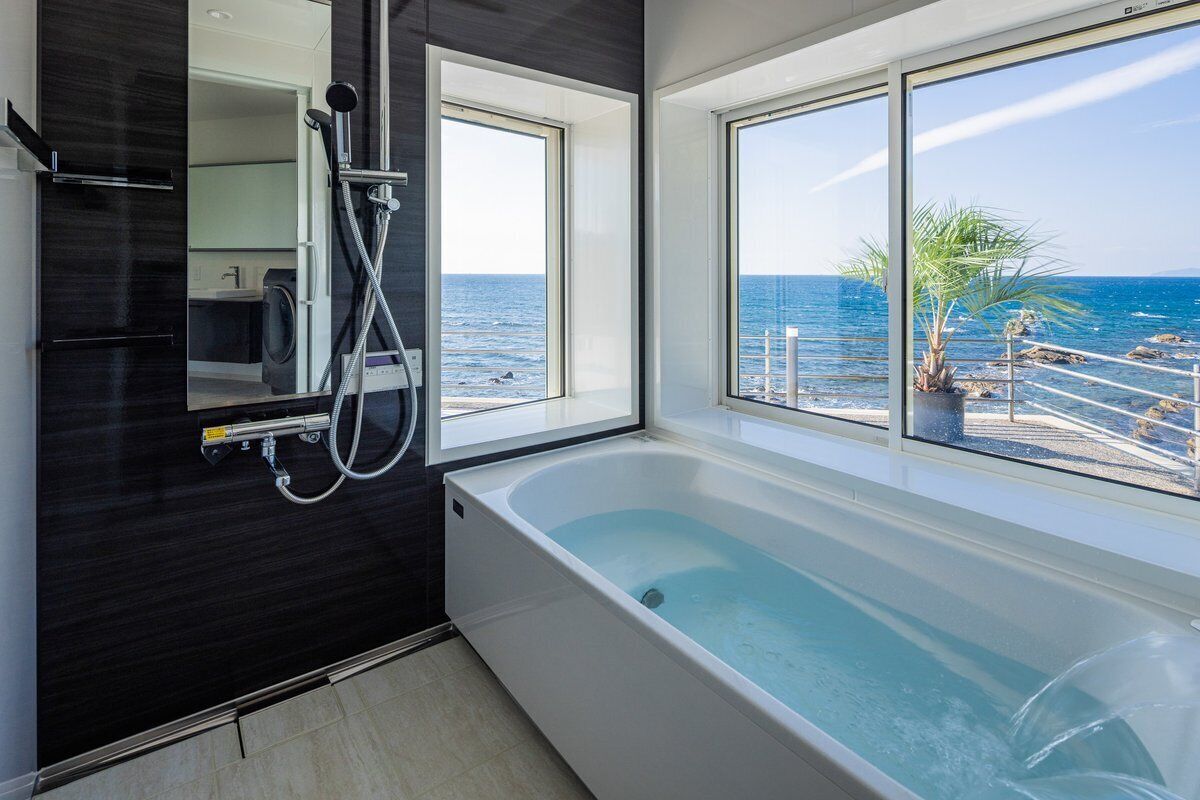 A modern bathroom with a bathtub, a shower, and a large window overlooking the sea.