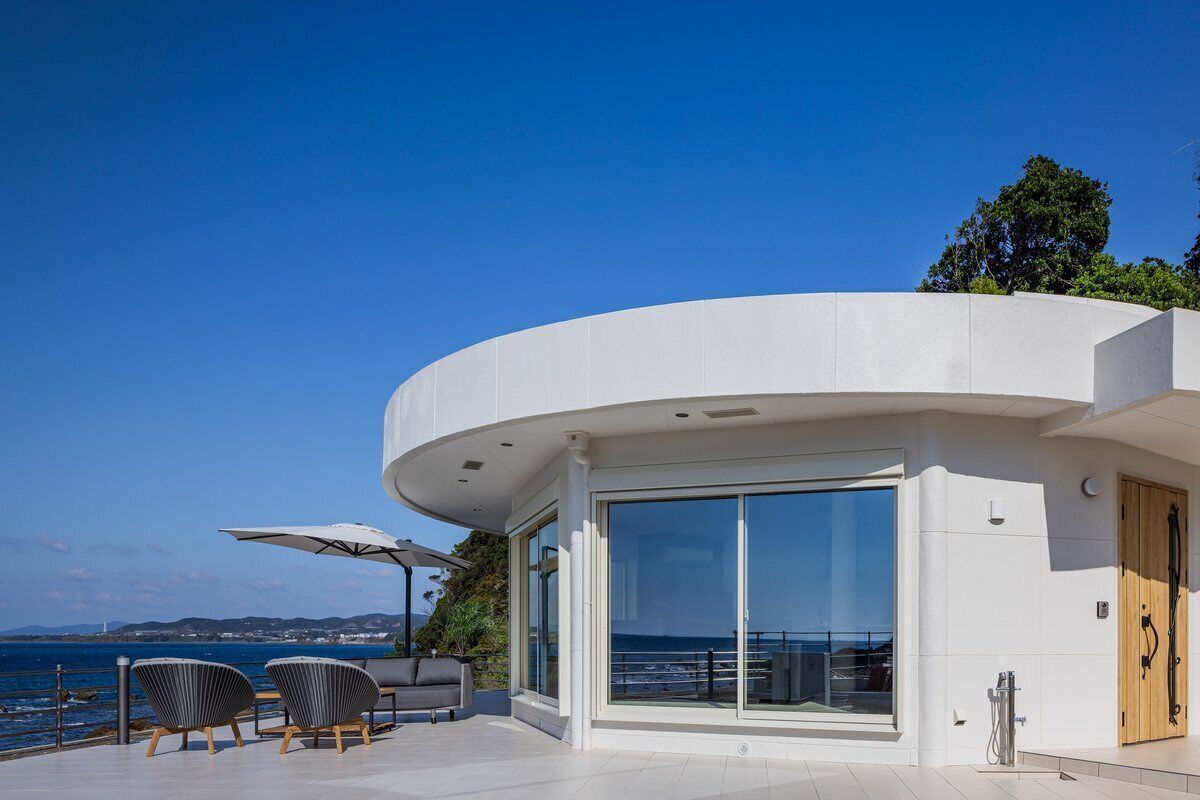 A modern building with a curved facade, large sliding glass doors, outdoor seating, and a view of the sea.