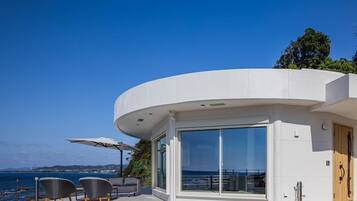 A modern building with a curved facade, large sliding glass doors, outdoor seating, and a view of the sea.