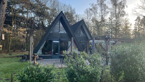 Een modern, driehoekig huis met grote ramen en een veranda, omgeven door weelderige begroeiing en bomen.