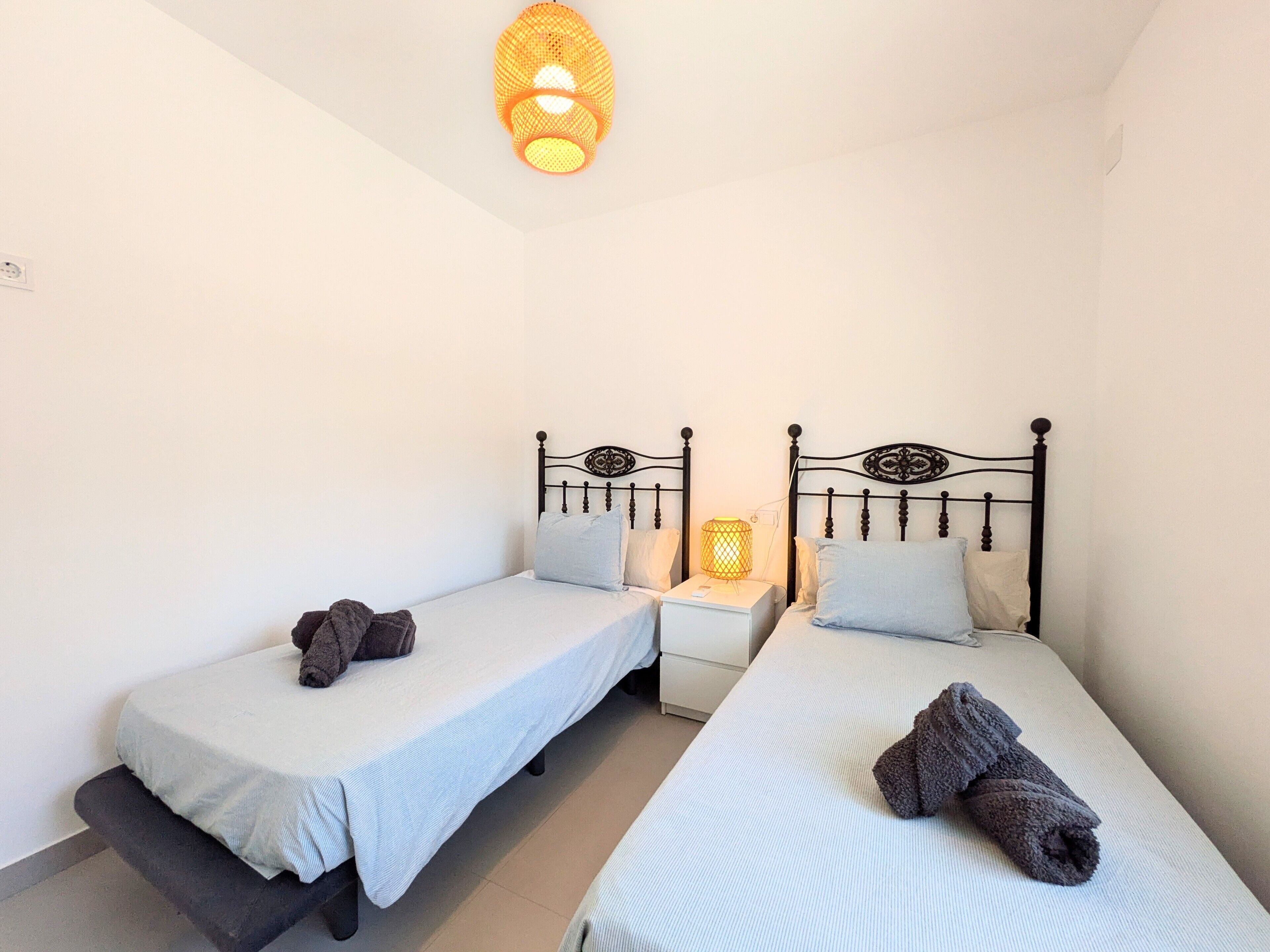 Una habitación pequeña y limpia con dos camas individuales, una mesita de noche con lámpara, y una lámpara fijada a la pared.