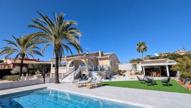 Un área de piscina con una casa, palmeras y muebles de exterior.