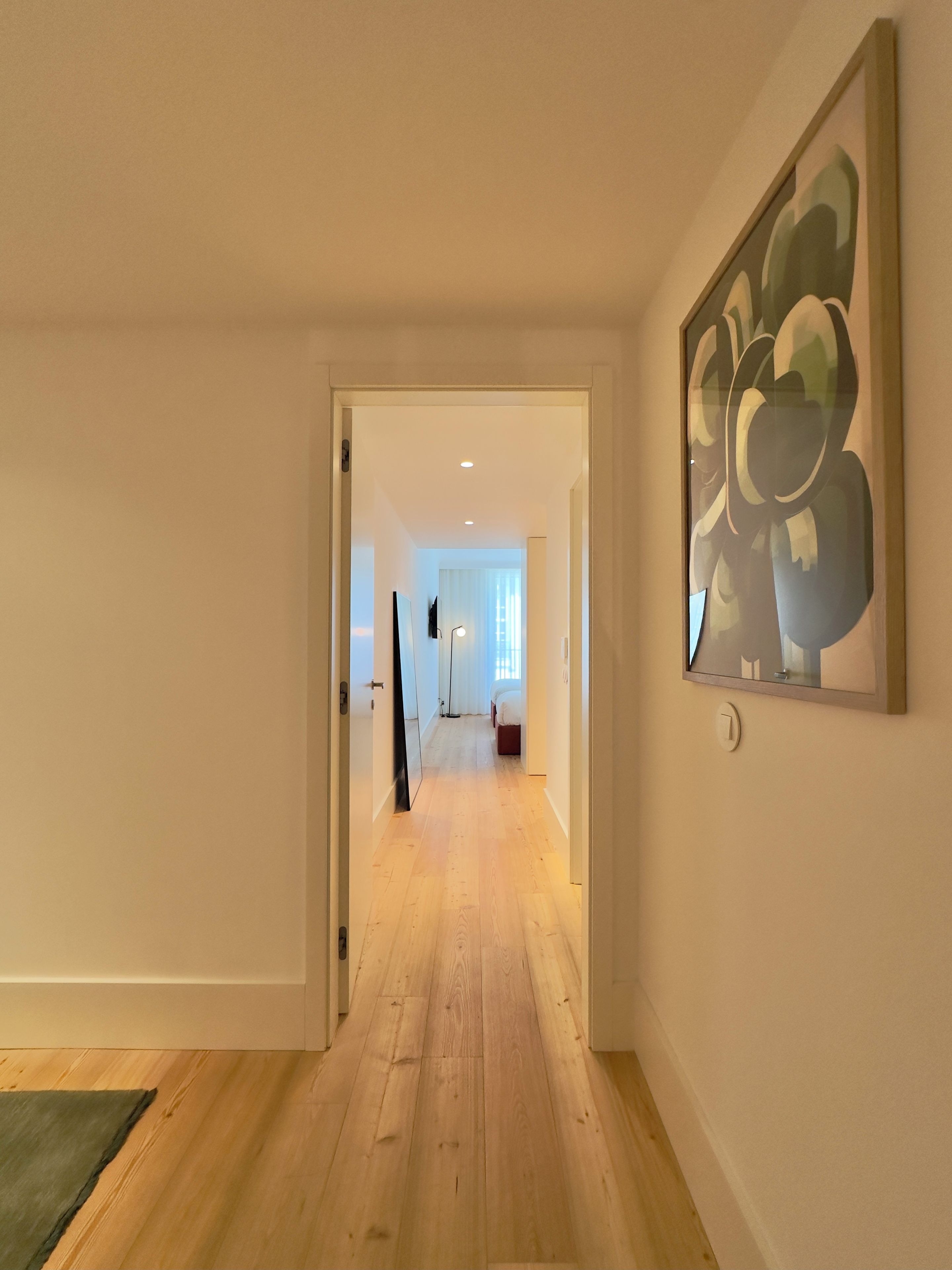 Un couloir au plancher de bois mène à une chambre à coucher qui contient un lit et une fenêtre.