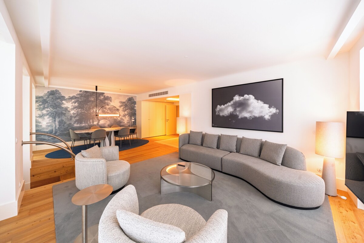 A modern living room with a curved sofa, round coffee table, and grey armchairs. A dining area is visible in the background with a chandelier.