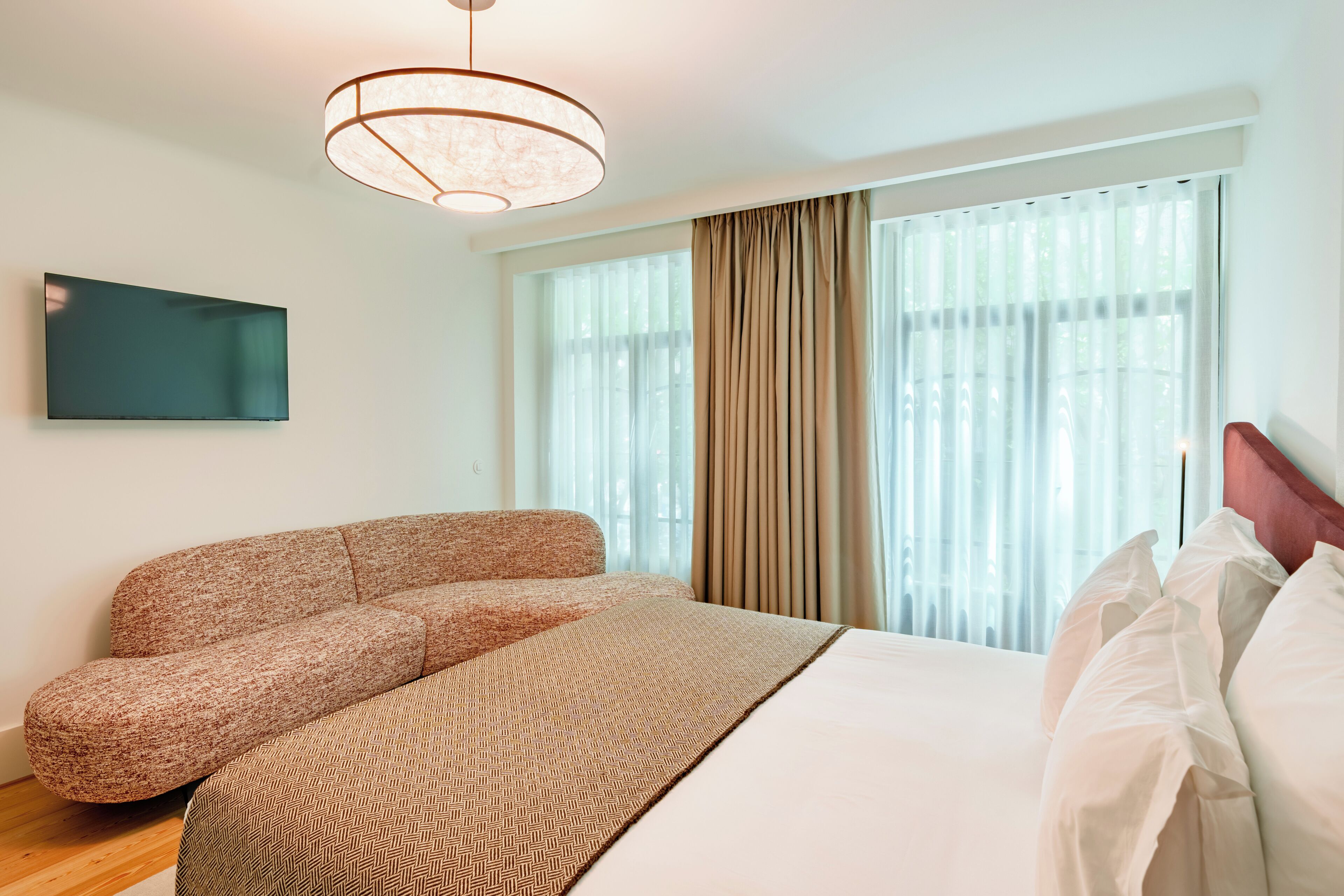 A modern hotel room with a bed, a sofa, a television, and a large window with curtains.