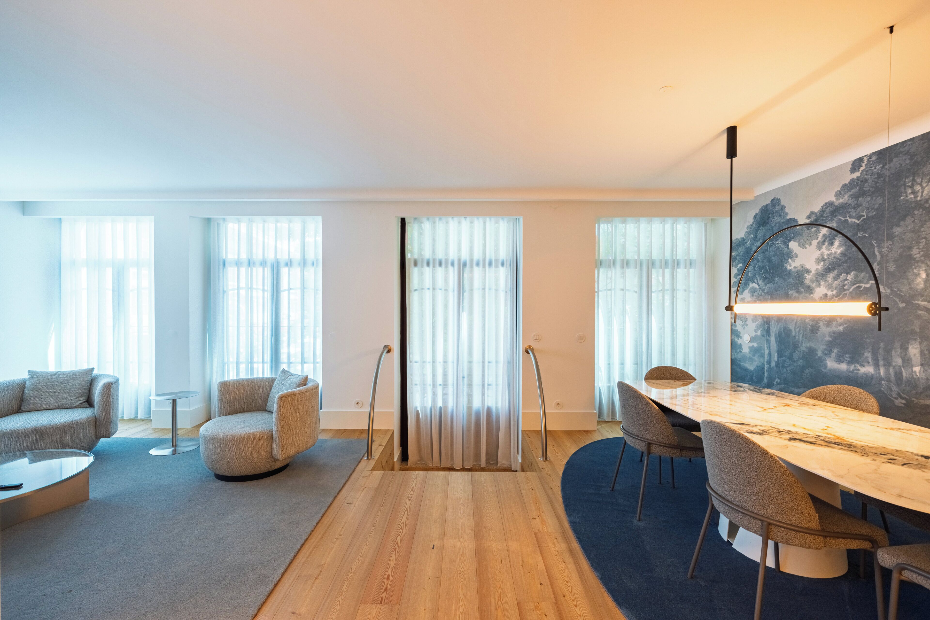 A modern living room with a wooden floor, a round coffee table, a sofa, a dining table with chairs, and a large window with curtains.