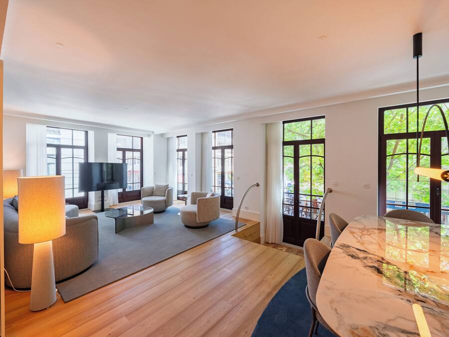Amplia sala de estar con televisor grande, asientos cómodos, zona de comedor con mesa de mármol y ventanas amplias con vistas.