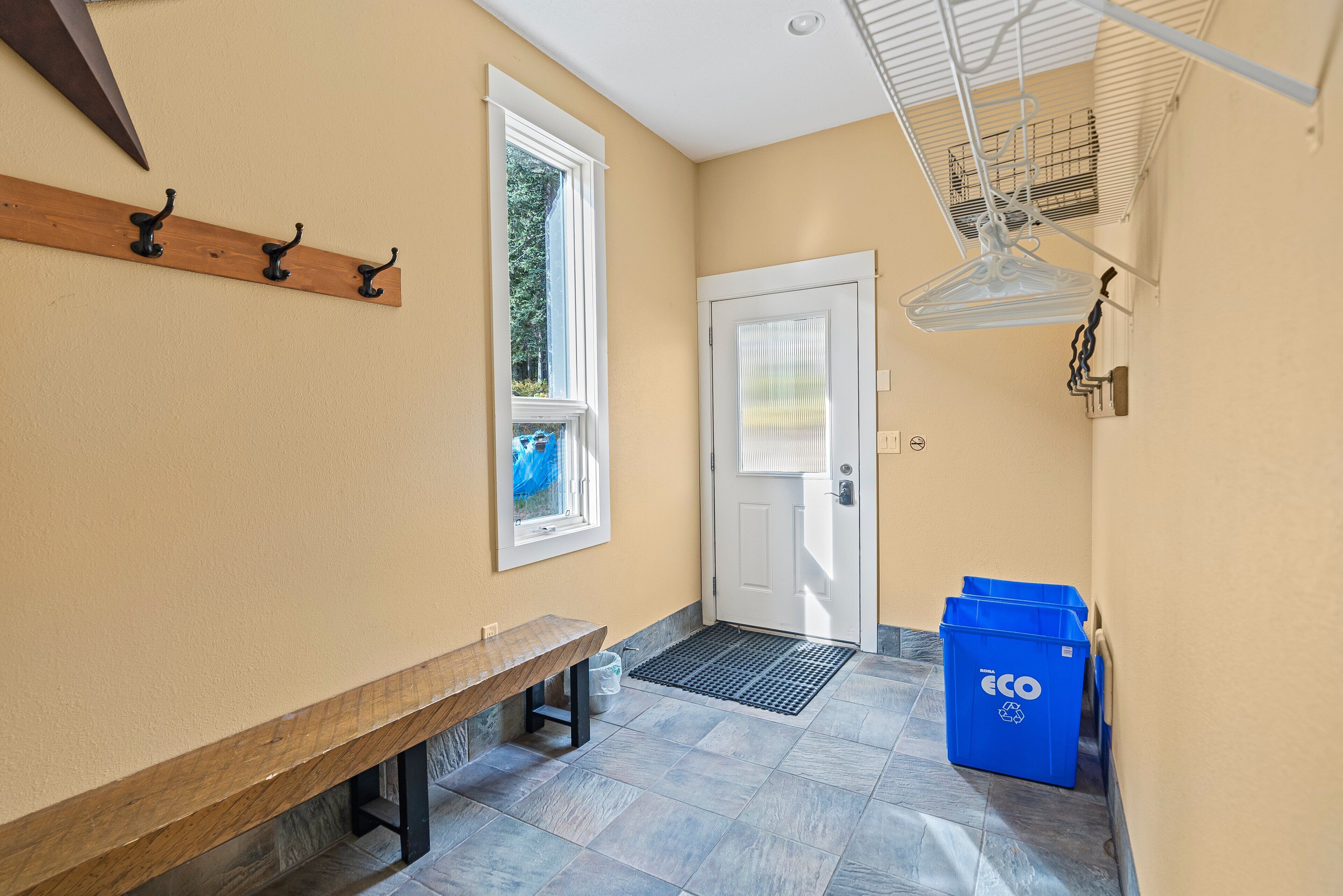 A hallway with a bench, a door, a window, and a blue recycling bin.