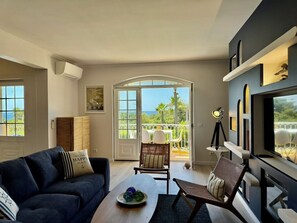 Ein Wohnzimmer mit einer Couch, Sesseln, einem Couchtisch, einem Fernseher und einem Balkon mit Aussicht.