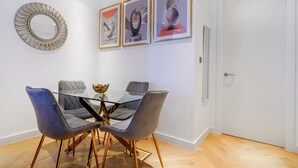 A dining area with a glass table and four grey upholstered chairs, a decorative mirror, and three framed artworks on the wall. - Cosy London Apartment With 2 Bedrooms (London)