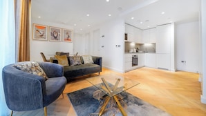 A modern living room with a sofa, a glass coffee table, and a kitchen area with white cabinets and a countertop. - Cosy London Apartment With 2 Bedrooms (London)