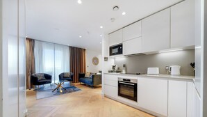 A modern kitchen with white cabinets, a microwave, and a coffee maker. There is a dining area with a blue sofa and a rug. - Cosy London Apartment With 2 Bedrooms (London)