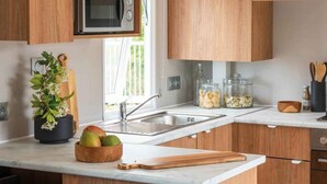 Une cuisine moderne avec des meubles en bois, un four à micro-ondes encastré, un évier et un plan de travail sur lequel se trouvent un saladier rempli de fruits et un rouleau à pâtisserie.