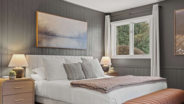 A bedroom with a large bed, two bedside tables with lamps, a bench, a window with curtains, and a painting on the wall. - Private Pool - Hot Tub - Game Room - 20 min DT (Sun Prairie)