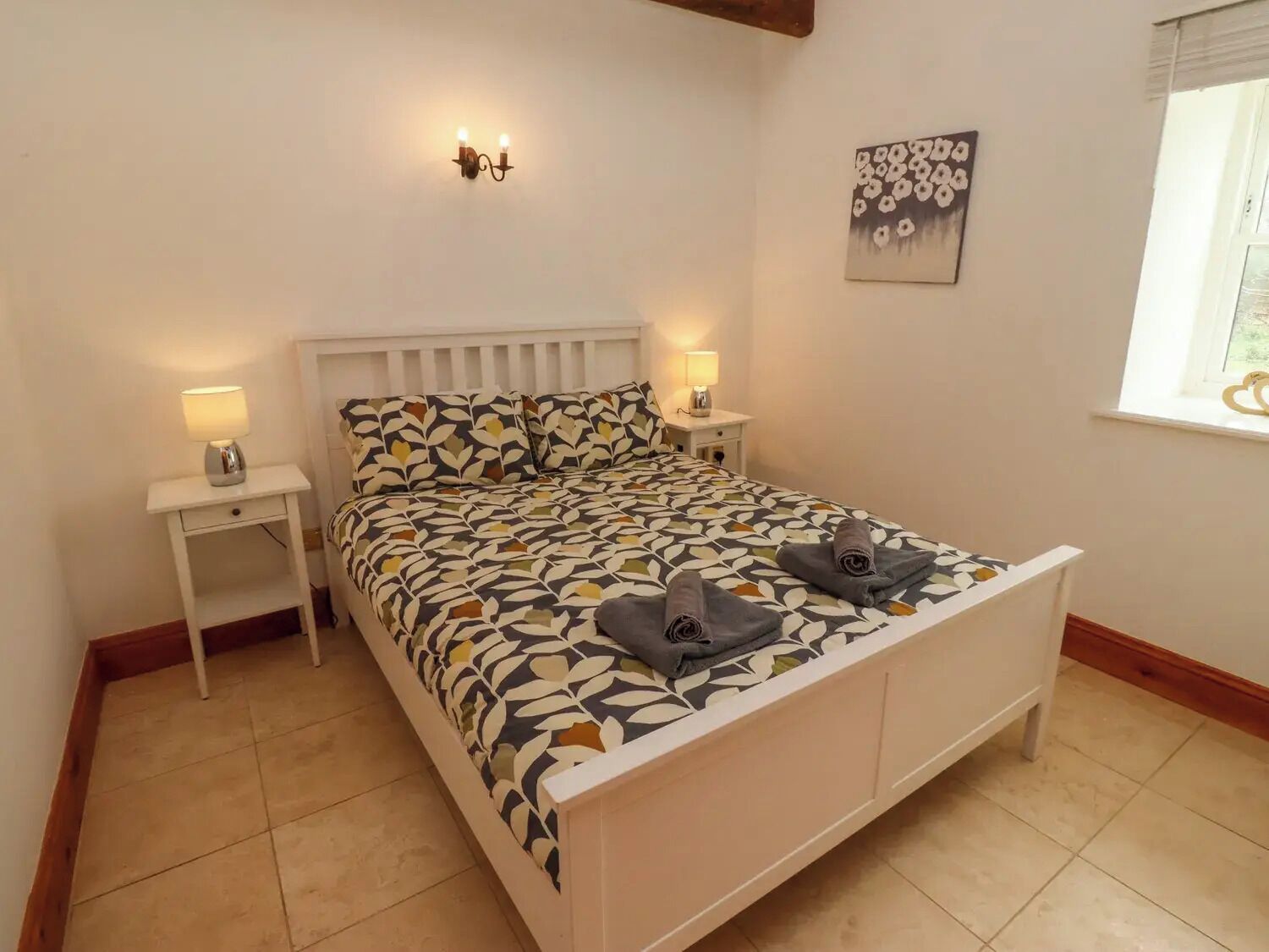 A neatly arranged bedroom with a patterned bedspread, bedside lamps, a wall-mounted light fixture, and a framed artwork.