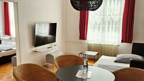 Ein modernes Zimmer mit Flachbildfernseher an der Wand, einem runden Tisch mit Stühlen, einem Bett mit weißem Kopfteil und roten Vorhängen.