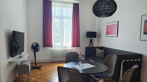 Ein modernes Wohnzimmer mit einem runden Esstisch, Korbstühlen, einem schwarzen Sofa, einem Fernseher, einem Ventilator und einem großen Fenster mit Vorhängen.