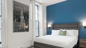A bedroom with a bed, nightstands, a wall-mounted lamp, a window with curtains, and a cityscape painting. - Olde City Loft Style Condo (Philadelphia)