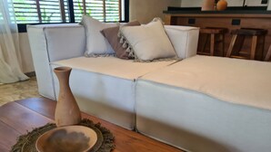 Una sala de estar con un sofá blanco, una mesa de centro de madera con un cuenco, y una cocina al fondo.
